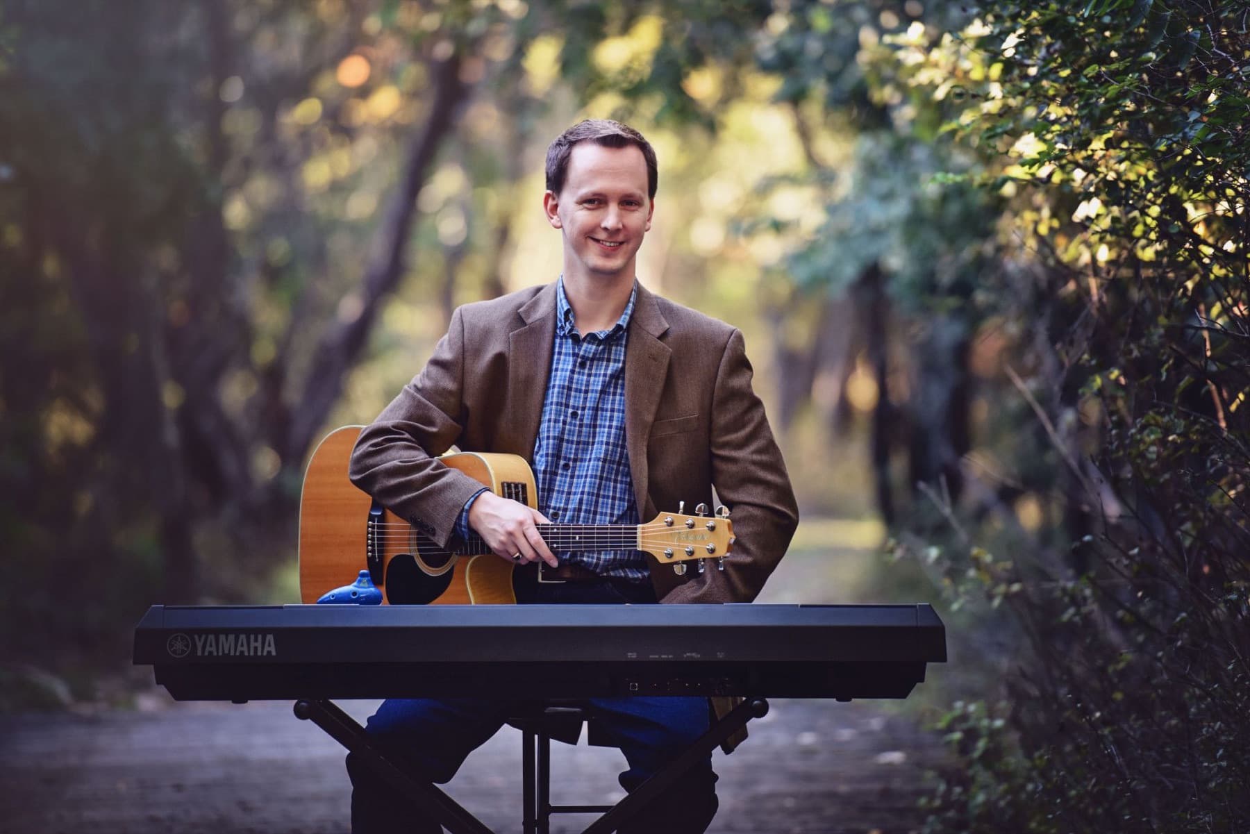 Stephen Forester with his guitar at a keyboard on a tree-lined path